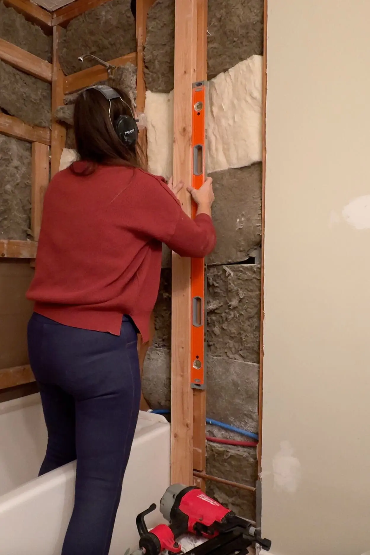 A person wearing ear protection and a red sweater is using a bright orange level to align a wooden vertical stud. The exposed wall shows insulation and piping, with a nail gun and other tools nearby.